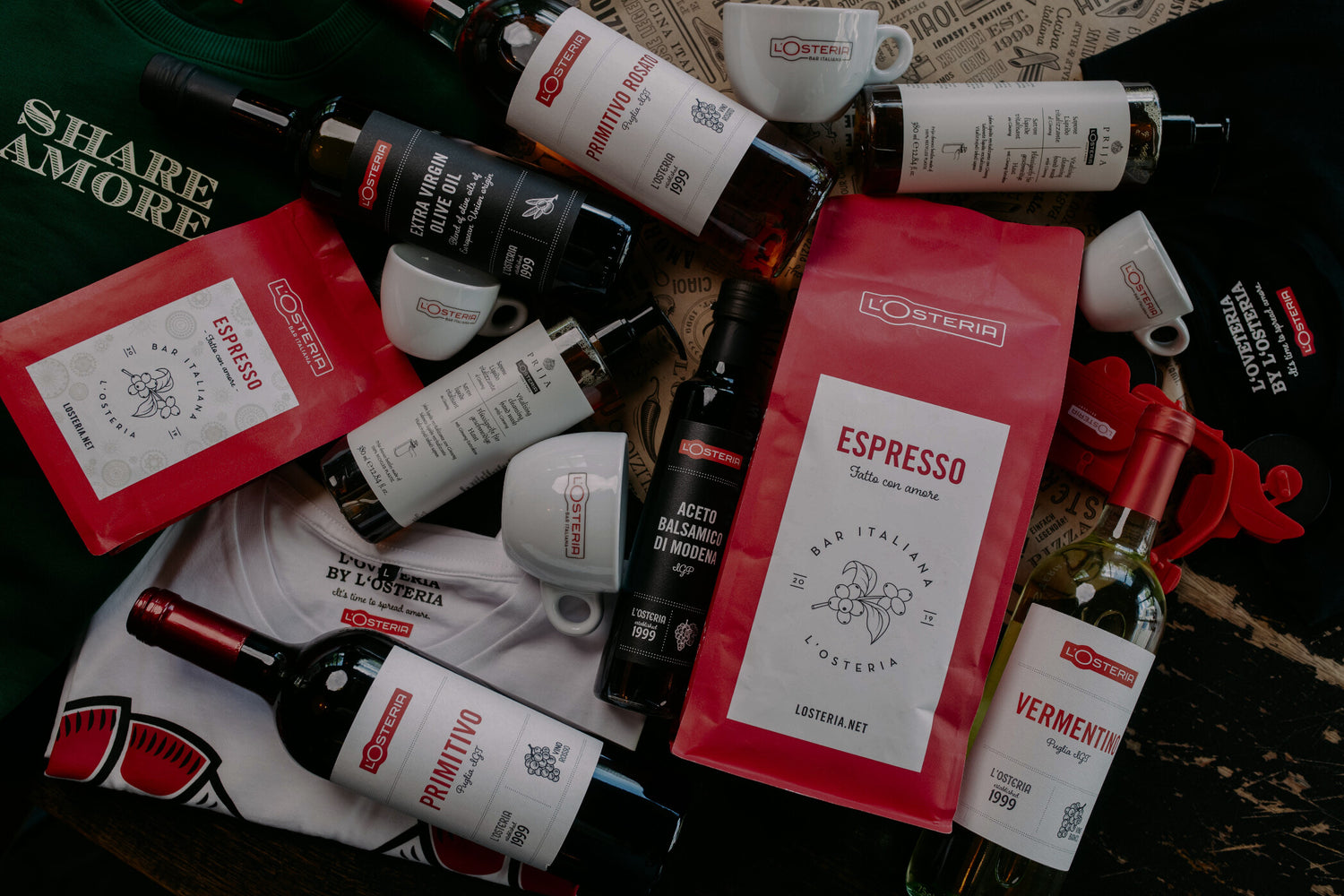L’Osteria Produkte wie Espresso, Wein, Olivenöl, Balsamico und Tassen liegen dekorativ arrangiert auf einem Tisch