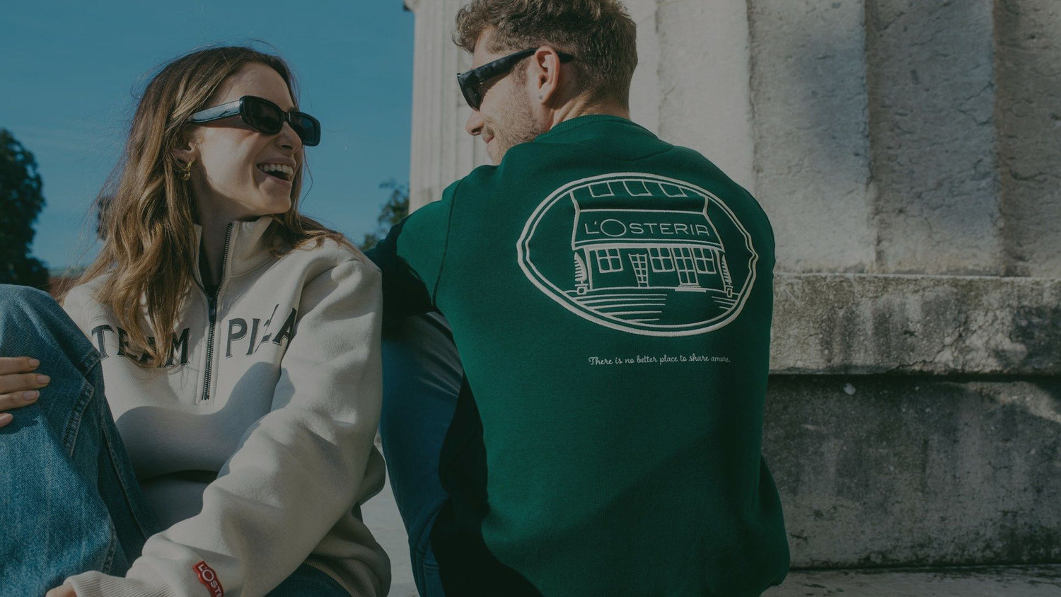 Zwei Personen tragen L’Osteria Pullover und sitzen entspannt im Freien, die Rückseite zeigt das Logo mit Restaurant-Illustration.